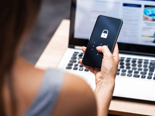Eine Frau sitzt vor einem Laptop und hat ein Handy mit einer Authenticator-App in der Hand