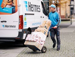 Ein Hermes Zusteller mit Paketen vor seinem Fahrzeug.