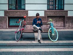 Junger Mann mit Handy, sitzt auf Parkbank, links und rechts ein sportlich modernes Fahrrad, Stadt Szenerie