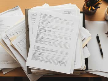 Ein Berg Akten liegt auf einem Schreibtisch. Ebenso findet sich dort eine Brille, ein Stift und eine leere Kaffeetasse.