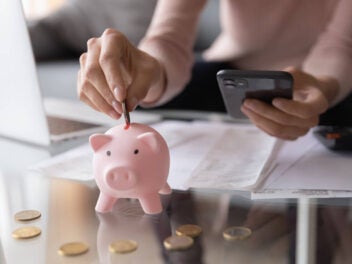 Frau mit Smartphone in der Hand wirft Geld in ein Sparschwein.