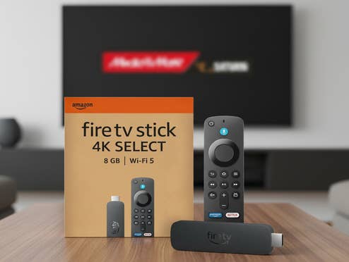 Der neue Amazon Fire TV Stick 4K Select steht auf einem Tisch in einem modernen Wohnzimmer. Im Hintergrund ist ein Fernseher zu erkennen, der das MediaMarkt/Saturn-Logo zeigt.
