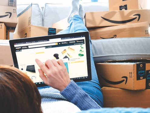 Wie viel sparst du wirklich mit deinem Amazon Prime Abo? Eine Frau liegt auf dem Boden, ihre Beine auf der Couch und sie tippt auf einem Tablet herum. Auf der Couch liegen mehrere Amazon-Pakete.