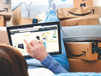 Eine Frau liegt auf dem Boden, ihre Beine auf der Couch und sie tippt auf einem Tablet herum. Auf der Couch liegen mehrere Amazon-Pakete.