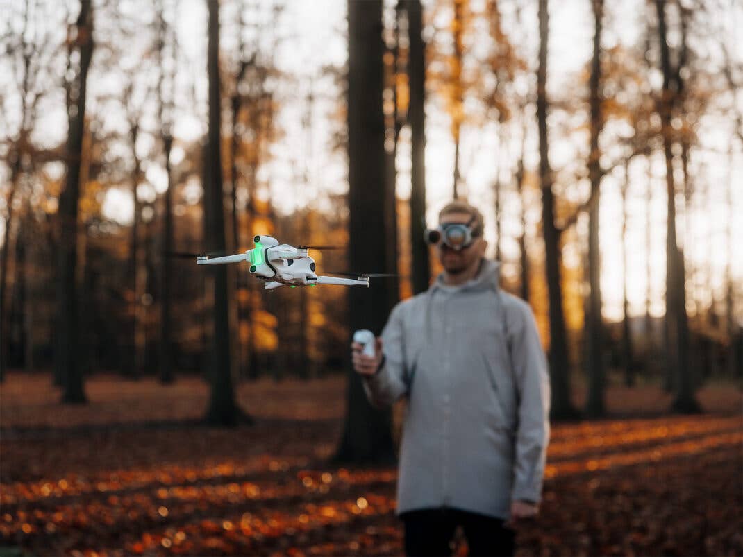 Drohne fliegt in einem Wald und wird von einem Mann im Hintergrund gesteuert.