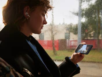 Person mit AirPods in den Ohren hält Apples iPhone SE 3 in einer Hand
