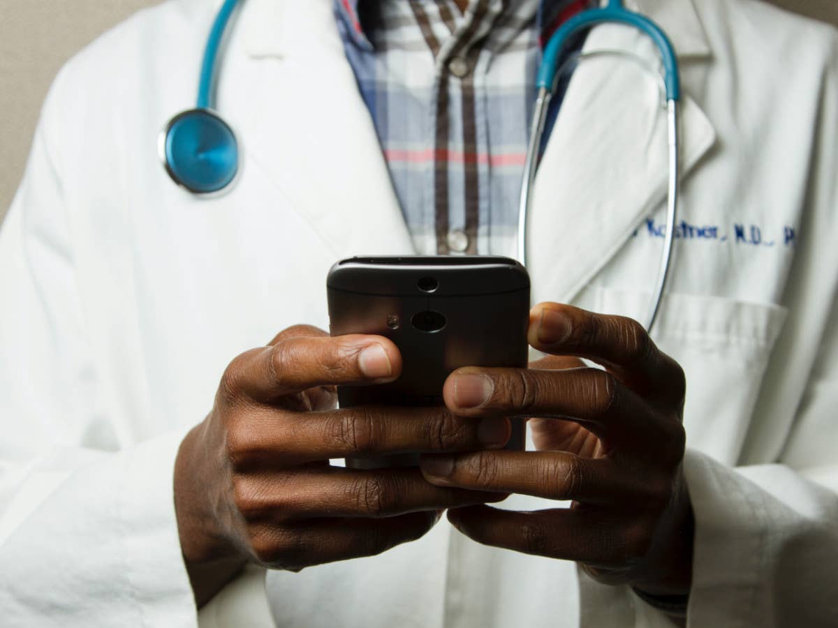 Ein Arzt hält ein Handy in der Hand, im Hintergrund sieht man seinen Kittel und ein Stethoskop.