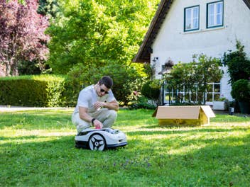 Person schaltet den Raccoon 2 SE Mähroboter per Knopfdruck im Garten an