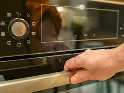 eine Hand will einen Backofen öffnen, man sieht die Symbole auf dem Gerät und den Drehregler