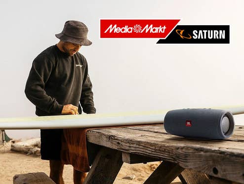 Surfer bearbeitet sein Surfboard, im Vordergrund rechts ein Bluetooth-Lautsprecher von JBL.