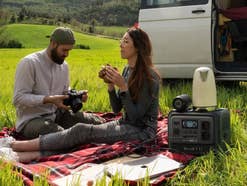 Zwei Personen sitzen auf einer Campingdecke vor einem Van in der Natur. Daneben steht die Bluetti Elite 100 V2.