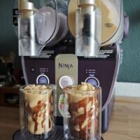Etwas Schokosoße dazu und fertig ist der Frappuccino. Ninja Slushi Twist mit beiger Mischung im rechten Tank, steht auf einer Küchentheke. Davor zwei Gläse mit Frappuccino und Schokosoße