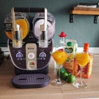 Die Frozen Margarita gelingt im Nu. Ninja Slushi Twist steht auf einer Küchentheke, im linken Tank ist eine orangene Flüssigkeit. Daneben stehen Flaschen und eine Saftpackung sowie zwei Martinigläser mit orangenem Slush gefüllt.