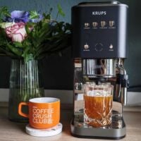 Die Krups Coffee Crush kann viel: Espresso, Cold Brew, Filterkaffee – aber schmeckt der auch gut? Krups Coffee Crush Experience Espresso-Vollautomat nebst Blumenwase und Dosen mit Tasse Kaffee und Glas mit Cold Brew