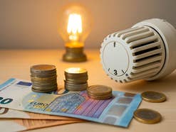 Geldscheine liegen auf einem Tisch, zusammen mit einem Heizungsthermostat und einer Glühbirne