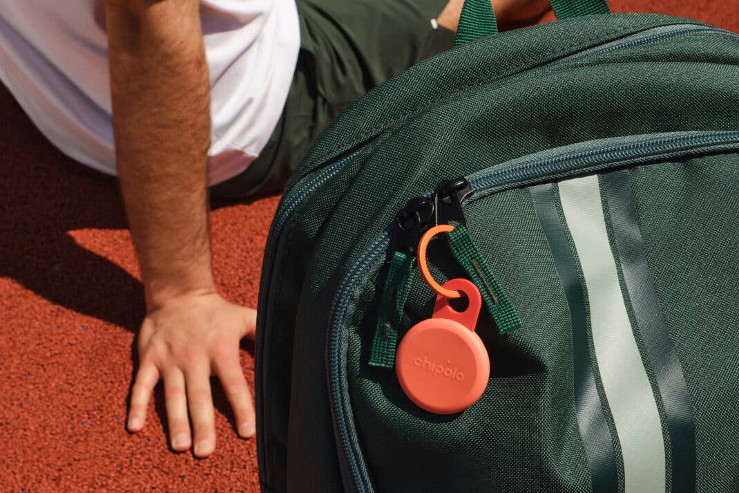 Ein Chipolo Loop-Artikel-Tracker, der an einem grünen Rucksack auf einer roten Fläche befestigt ist.