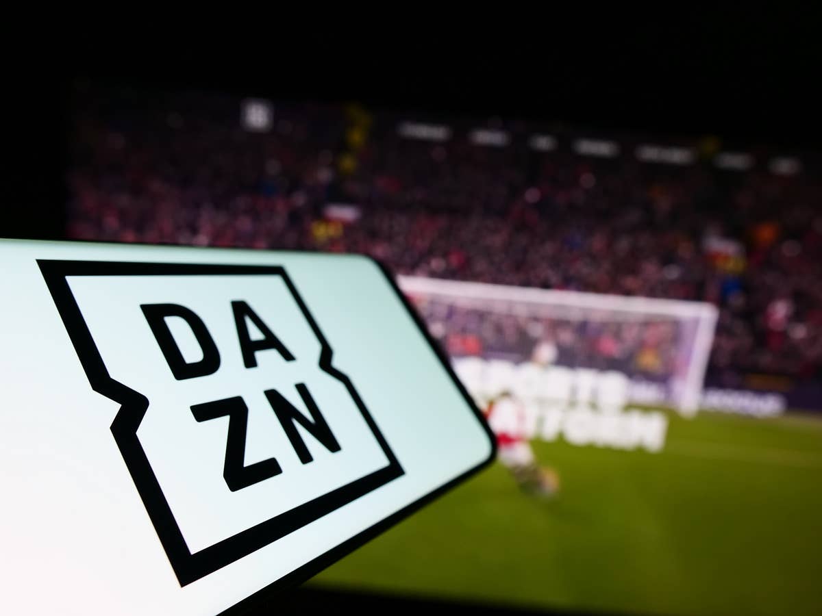 DAZN-Logo auf einem Smartphone vor einem Fernseher im Hintergrund, auf dem ein Fußballspiel läuft.