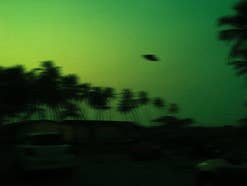 Ein Ufo ist an einem grünen Himmel zu sehen; im Hintergrund Palmen.