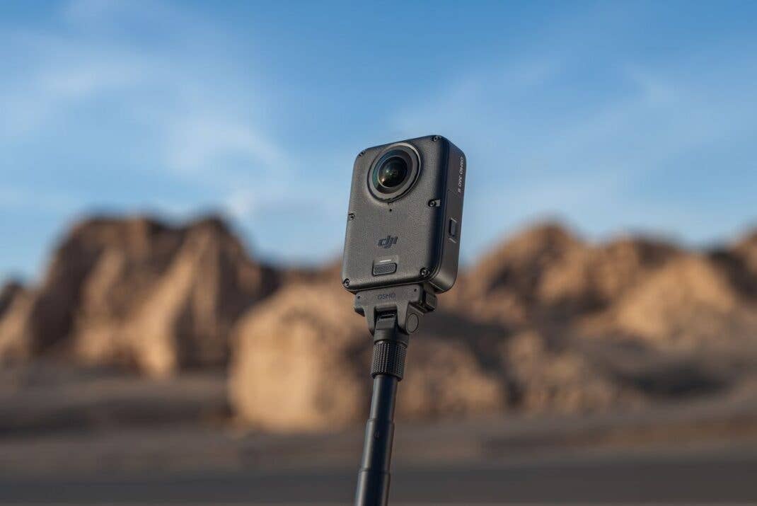 Die schwarze DJI-Osmo-360-II-Kamera steckt auf einem ausgefahrenen Selfie-Stick vor unscharfem Hintergrund mit Felsformationen und blauem Himmel.