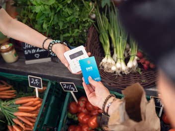 Eine Person zahlt mit einer Karte. Eine andere Person hält das Bezahlterminal. Beide sind in einem Gemüseladen.