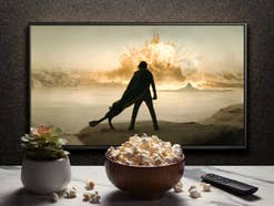 Bildschirm zeigt dunkle Figur in der Wüste vor einer Explosion, davor ein Tisch mit Pflanze, Popcorn und Fernbediehnung