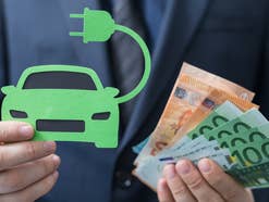 Mann hält ein Symbolbild für ein E-Auto und Euro-Geldnoten in der Hand.