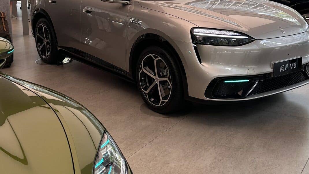 Ein beiges Elektro-SUV mit Leuchtstreifen an der Front steht in einem Showroom neben weiteren Fahrzeugen.