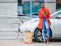 Frau lädt ihr Elektroauto beim Einkaufen und nutzt ein Smartphone.