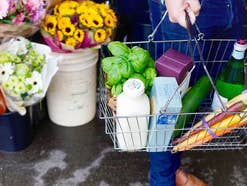 Eine Person hält einen Einkaufskorb, in dem Dinge liegen, im Hintergrund sind Blumen.