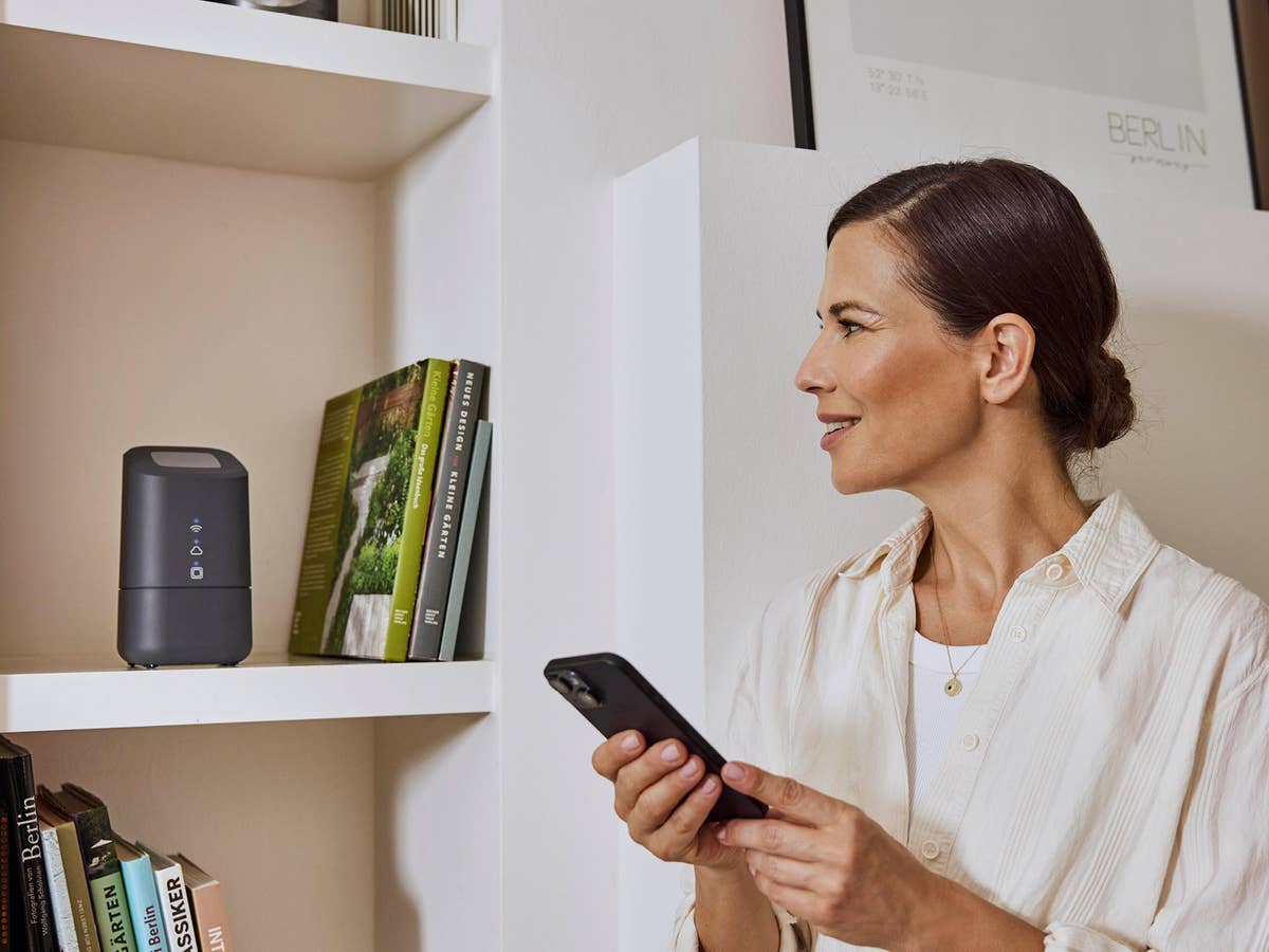 Homematic IP startet mit neuer Home Control Unit durch Eine Frau bedient per App die Home Control Unit, die hinter ihr im Regal steht.