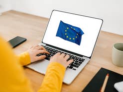 Frau arbeitet am Laptop, mit Kaffe-Tasse, Handy, Notizbuch, Stift und Kopfhörerladeetui daneben. Auf dem aufgeklappten Bildschirm ist eine EU-Fahne zu sehen.