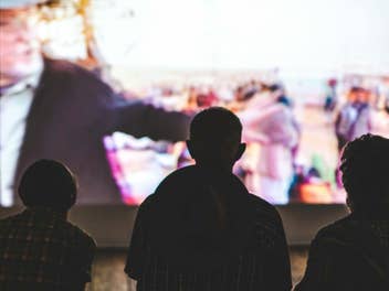 Drei Menschen sitzen vor einer großen Leinwand, auf der ein Film läuft.