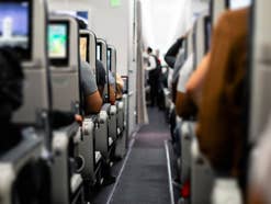 Flugzeug von innen, mehrere Sitzplätze sind belegt und Bildschirme laufen