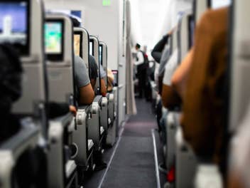 Flugzeug von innen, mehrere Sitzplätze sind belegt und Bildschirme laufen