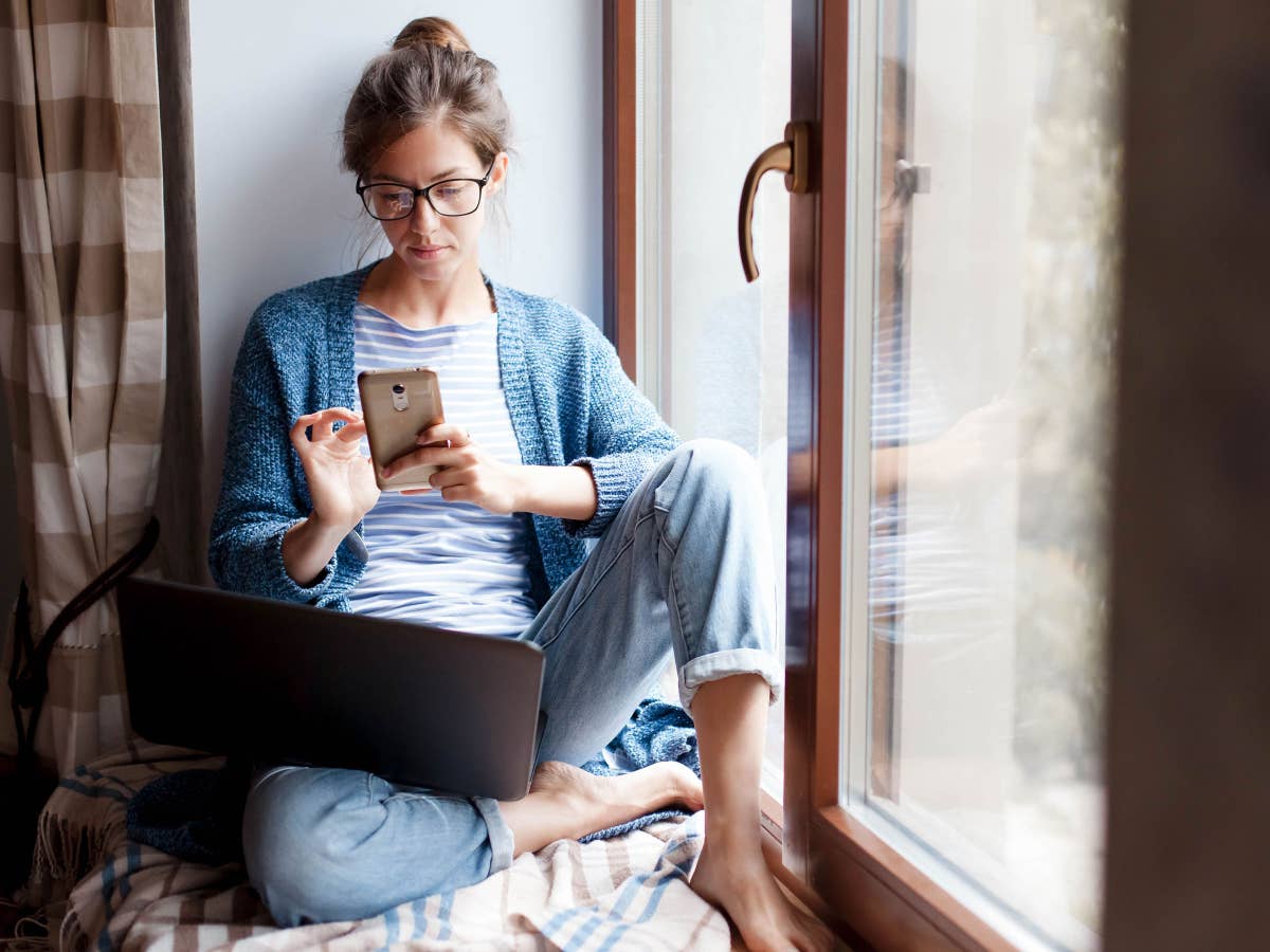 Frau sitzt mit Smartphone und Notebook am Fenster