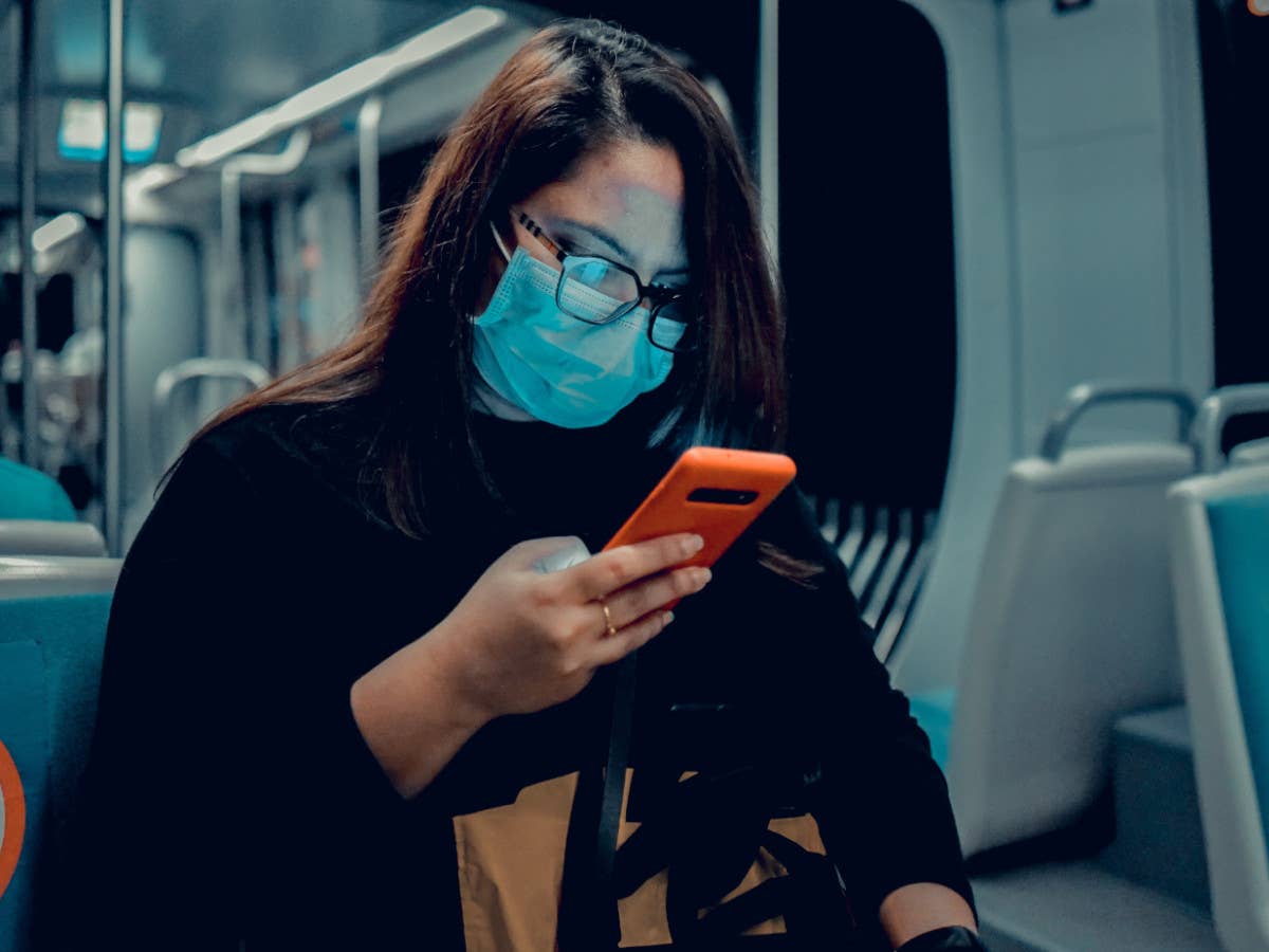 Eine Frau mit OP-Maske sitzt in einer U-Bahn und schaut auf ihr Handy.