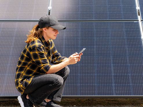 Eine Frau hockt vor einer Solaranlage mit einem Handy in der Hand