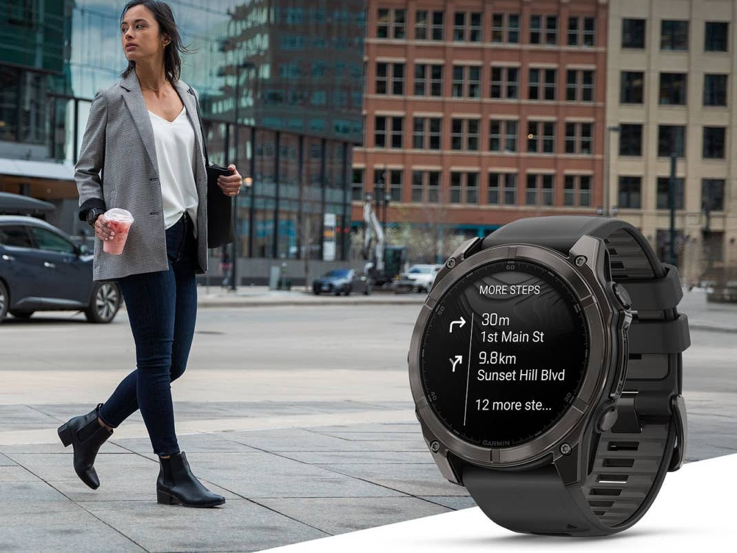 Eine Frau, die in einer Stadt geht, trägt einen Blazer, hält ein Getränk und hat eine Garmin-Uhr, die Wegbeschreibungen anzeigt.