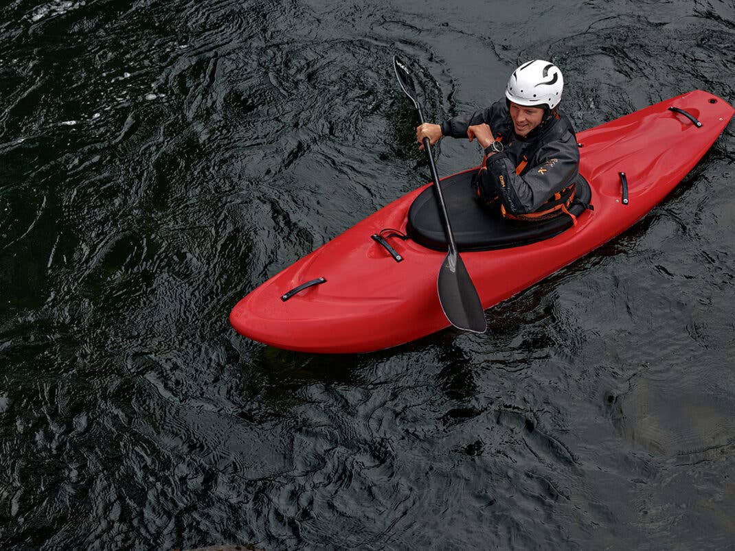 Eine Person paddelt in einem roten Kajak auf dunklem Wasser und trägt einen Helm und einen Neoprenanzug.