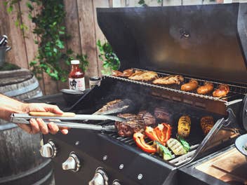 Eine Person dreht Grillgut auf dem Rost eines aufgeklappten Gasgrills