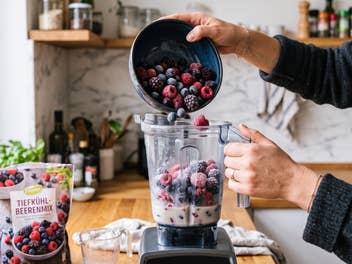 Eine Frau befüllt einen Standmixer mit Tiefkühlfrüchten in ihrer Küche