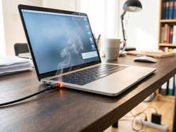 Ein Laptop, der geladen wird, doch das Ladekabel fängt an der Buchse an zu qualmen. Der Laptop steht auf einem Schreibtisch mit Lampe, Tasse und Unterladen. Im Hintergrund sieht man noch ein Bücherregal und eine Mehrfachsteckdose auf dem Boden.