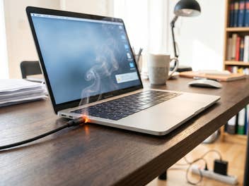 Ein Laptop, der geladen wird, doch das Ladekabel fängt an der Buchse an zu qualmen. Der Laptop steht auf einem Schreibtisch mit Lampe, Tasse und Unterladen. Im Hintergrund sieht man noch ein Bücherregal und eine Mehrfachsteckdose auf dem Boden.
