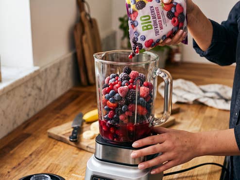 Eine Frau befüllt einen Standmixer mit Tiefkühlfrüchten in ihrer Küche