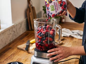 Eine Frau befüllt einen Standmixer mit Tiefkühlfrüchten in ihrer Küche