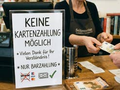 Mann zahlt an der Kasse mit Bargeld. Die Kassierein nimmt das Geld entgegen. Daneben steht ein Hinweisschild, dass dort nur mit Bargeld gezahlt werden kann.