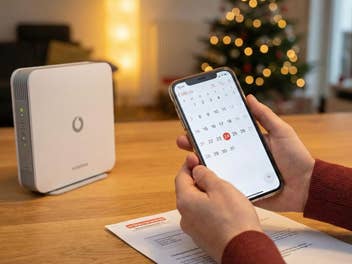 Ein Vodafone-Router auf einem TIsch, im Hintergrund ein Tannenbaum und jemand hält ein Smartphone in der Hand.