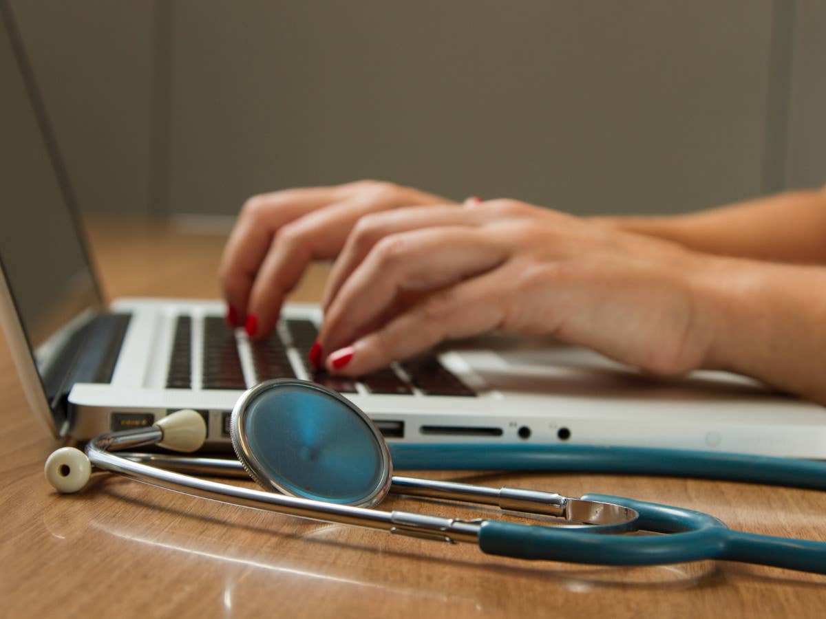 Ein Stethoskop liegt vor einem Laptop, auf dessen Tastatur eine Person tippt.