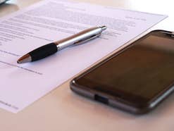 Symbolbild Handyvertrag: Ein Handy, ein beschriebenes Blatt Papier und ein Stift liegen auf einem Tisch