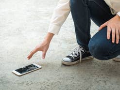 Person hebt ein zu Boden gefallenes Smartphone mit Displayschaden auf.